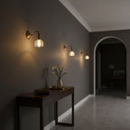 Three Clérachic brass wall lights run along a dark grey wall, above a narrow console table with a vase of flowers.