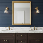 A pair of Clérachic wall lights with glass shades flank a gold-framed mirror on a navy boarded wall, above a double marble vanity.