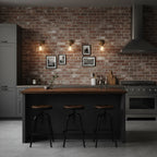 Three Clérachic wall lights glow on a red brick wall above a black kitchen island with round stools and framed prints.