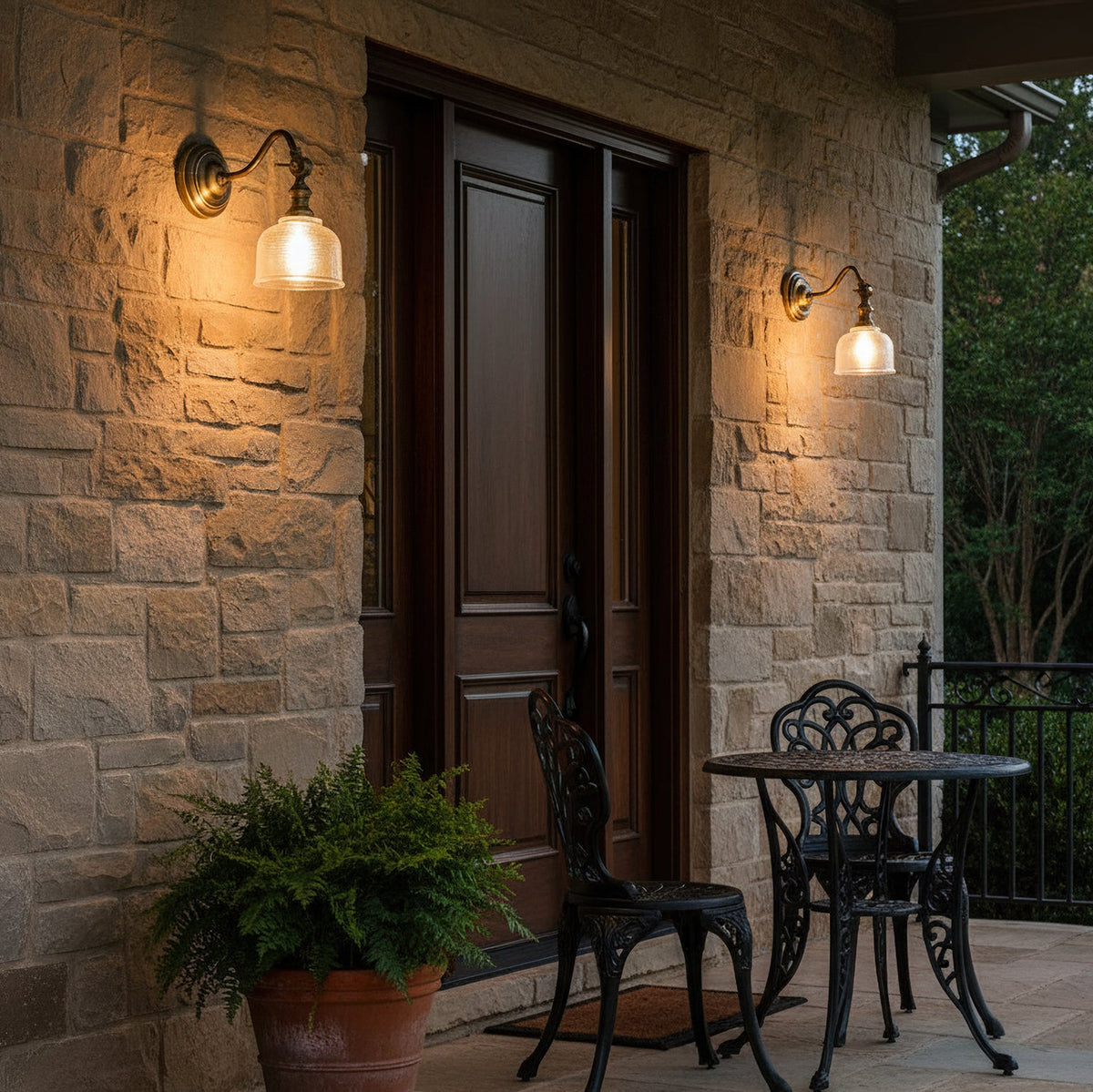 Two Clérachic wall lights with glass shades glow on a stone wall either side of a dark wooden front door, with a bistro set nearby.