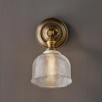 Close view of the ribbed glass shade, brass neck and round backplate of this Clérachic wall light, lit on a grey wall.