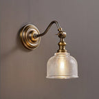 Side view of this Clérachic wall light with its curved brass arm, round backplate and ribbed clear glass shade, lit on a taupe wall.