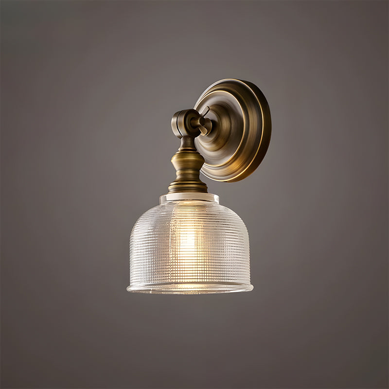 Clérachic wall light seen from the side, with a short brass arm, a round backplate and a ribbed glass shade on a brown-grey wall.