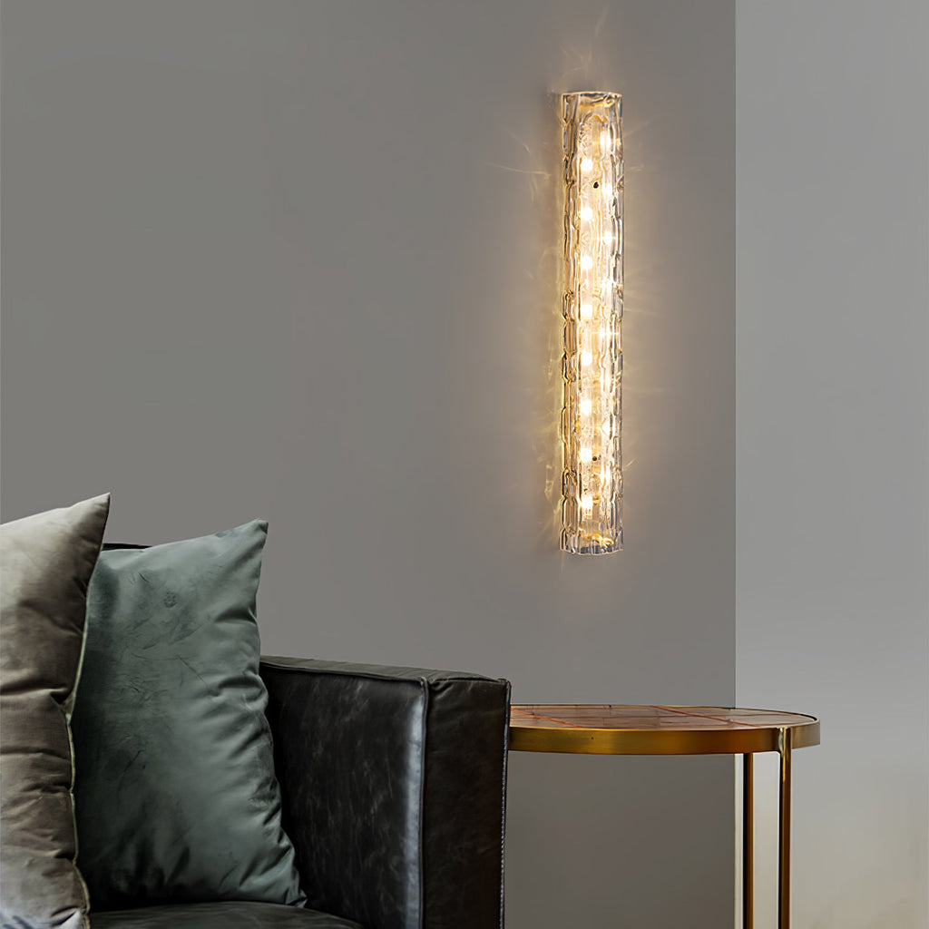Lit Clérachic wall sconce with a clear textured glass shade, mounted on a grey wall above a leather sofa and a round brass side table.