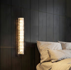 A Clérachic glass wall light glows on a dark wood-panelled wall, above a bed with beige pillows and linen.