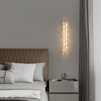 Slim Clérachic wall light lit beside a grey curtain, above a bedside cabinet and a bed with a tan quilted headboard.