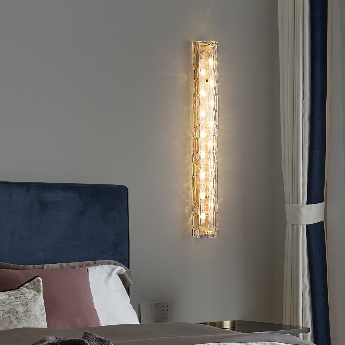 This Clérachic wall light glows beside a curtain edged in navy, above a bed with a blue velvet headboard and rose pillows.