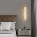 A Clérachic glass wall light lit above a bedside cabinet, beside a grey curtain and a tan headboard with a woven pattern.