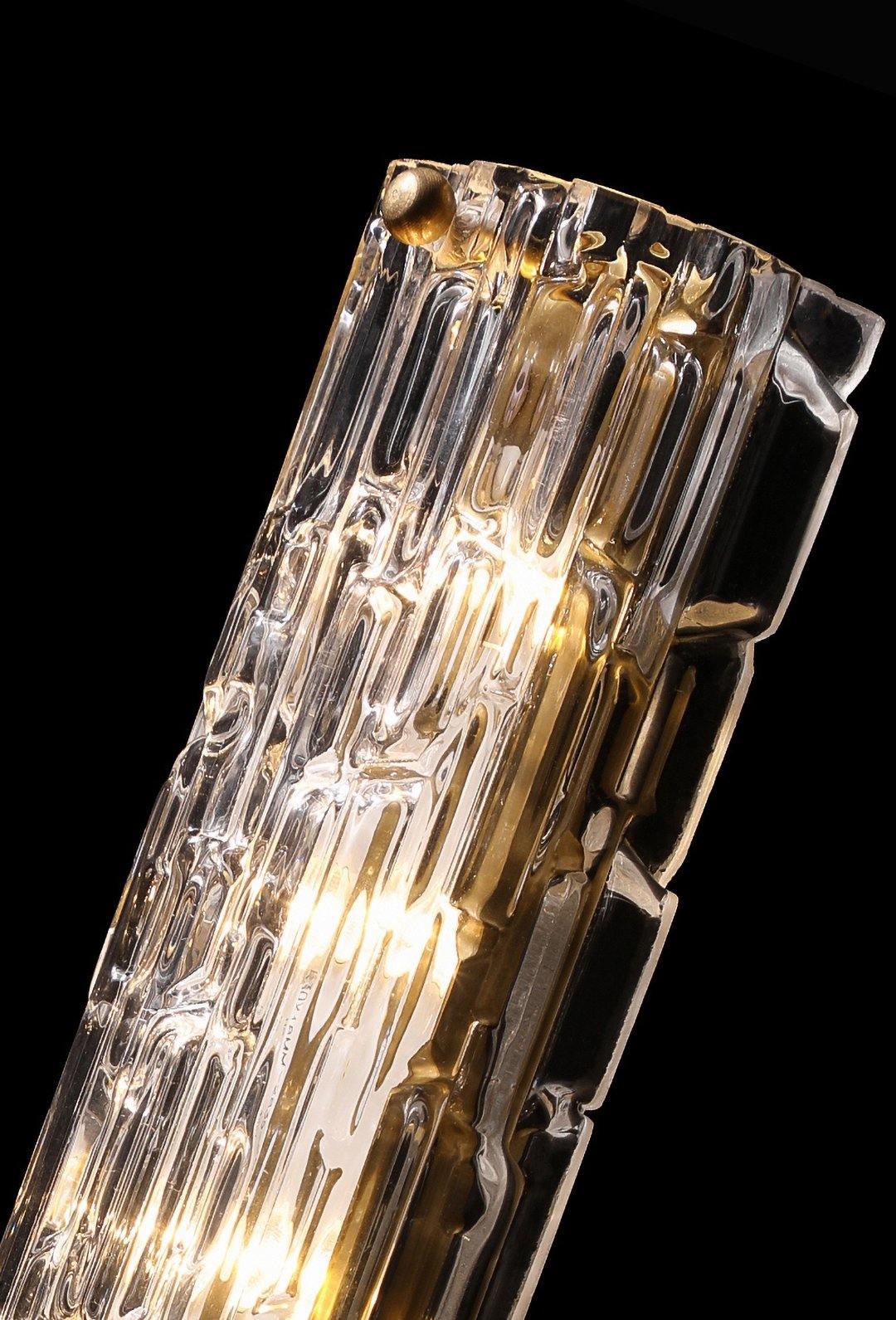 Close view of the clear, rippled glass of this Clérachic wall light, with the gold backplate and bulbs glowing inside, on black.