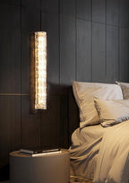 Clérachic glass sconce on a dark slatted wall, lit above a round bedside table with books and a bed in warm linen.