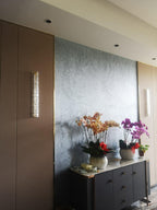 A Clérachic wall light, lit, on a brown panel beside a textured blue-grey wall, with orchids on a dark sideboard below.