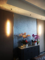 Clérachic wall light glowing as a bright strip on a brown panel, beside a blue-grey wall with orchids on a sideboard.