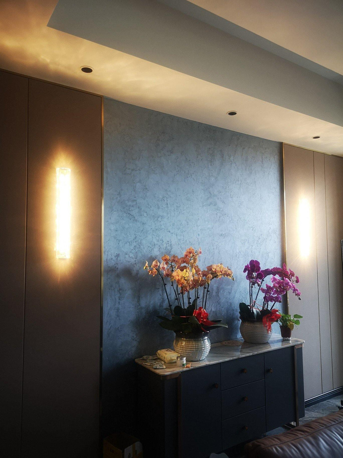 Clérachic wall light glowing as a bright strip on a brown panel, beside a blue-grey wall with orchids on a sideboard.