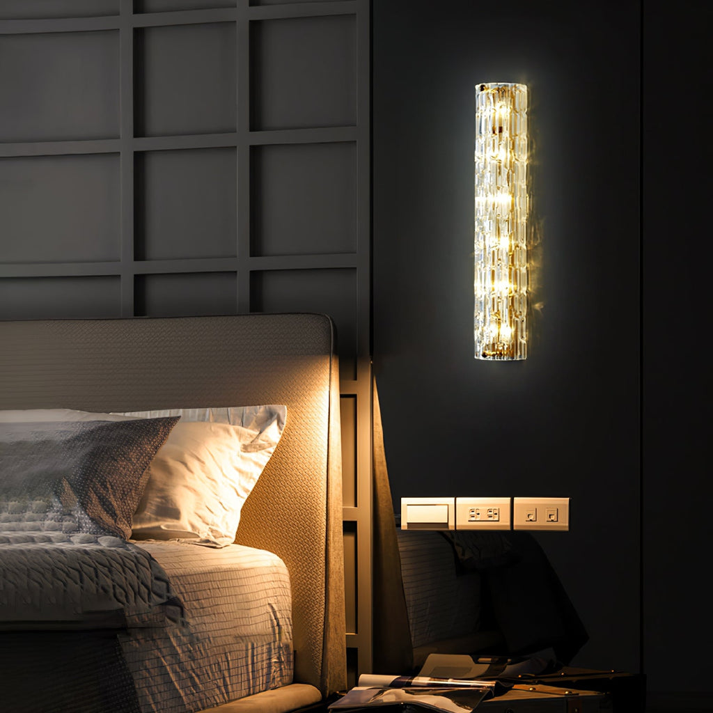 A Clérachic wall light lit on a dark panelled wall above a bedside shelf with sockets, with a bed glowing in warm light at the left.