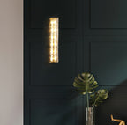 Clérachic wall light lit on a dark green panelled wall, above a glass vase with the leaves of a large plant.