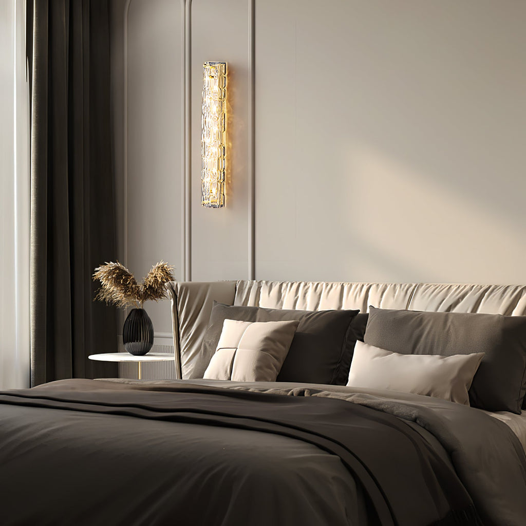 Clérachic wall light lit on a pale wall by a dark curtain, above a bed with grey linen, cushions and a dried-grass vase.