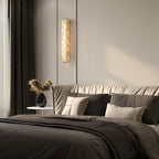 Clérachic wall light lit on a pale wall by a dark curtain, above a bed with grey linen, cushions and a dried-grass vase.