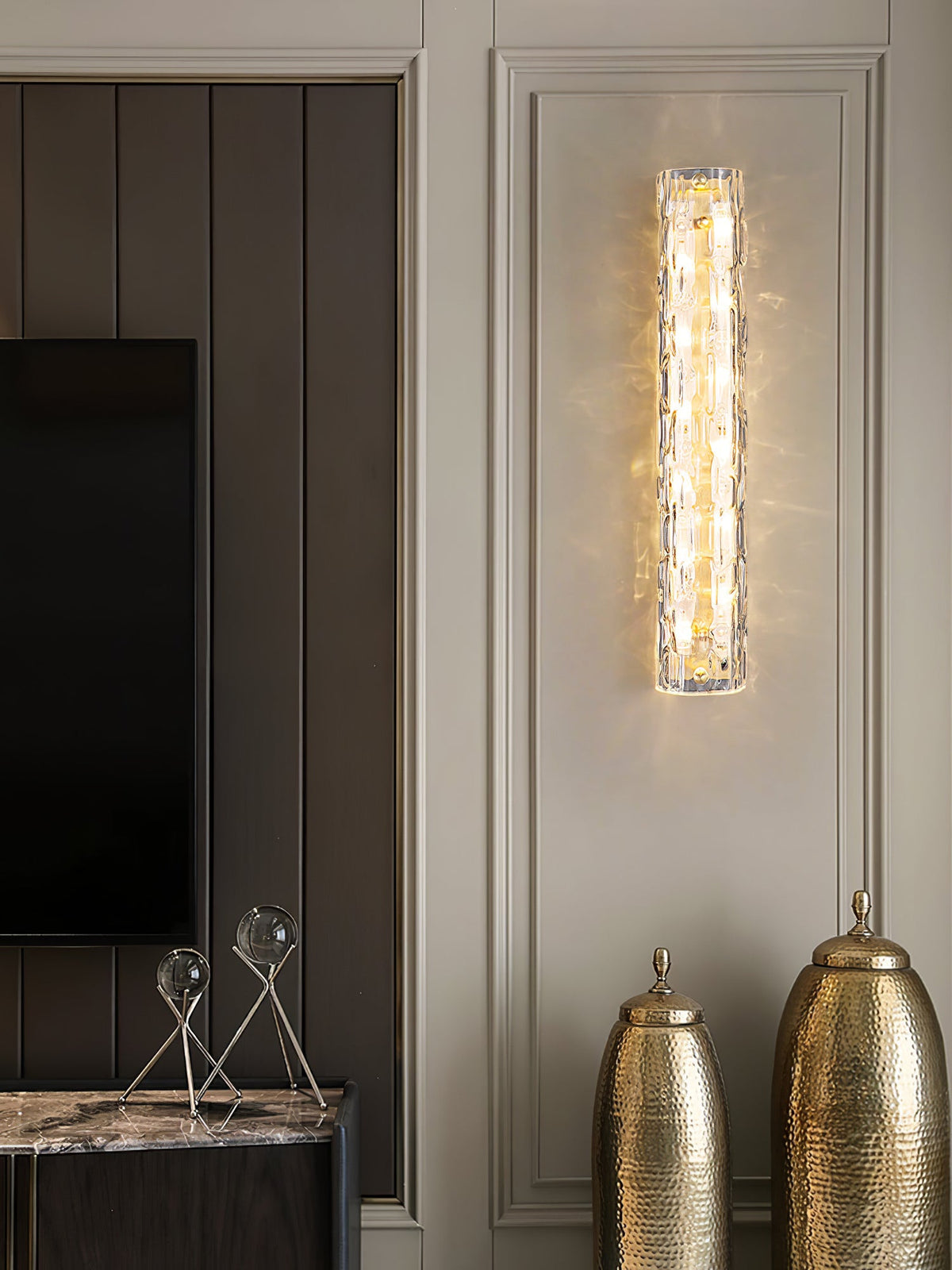 A Clérachic wall light glows on a panelled wall beside a dark TV, with two hammered gold urns and glass ornaments below.