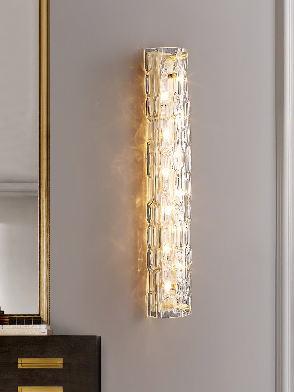 Close view of a lit Clérachic wall light on a grey wall beside a gold-framed mirror and a dark cabinet with brass handles.