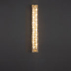 A taller Clérachic wall light glowing on a dark grey wall, its textured clear glass catching the warm light.