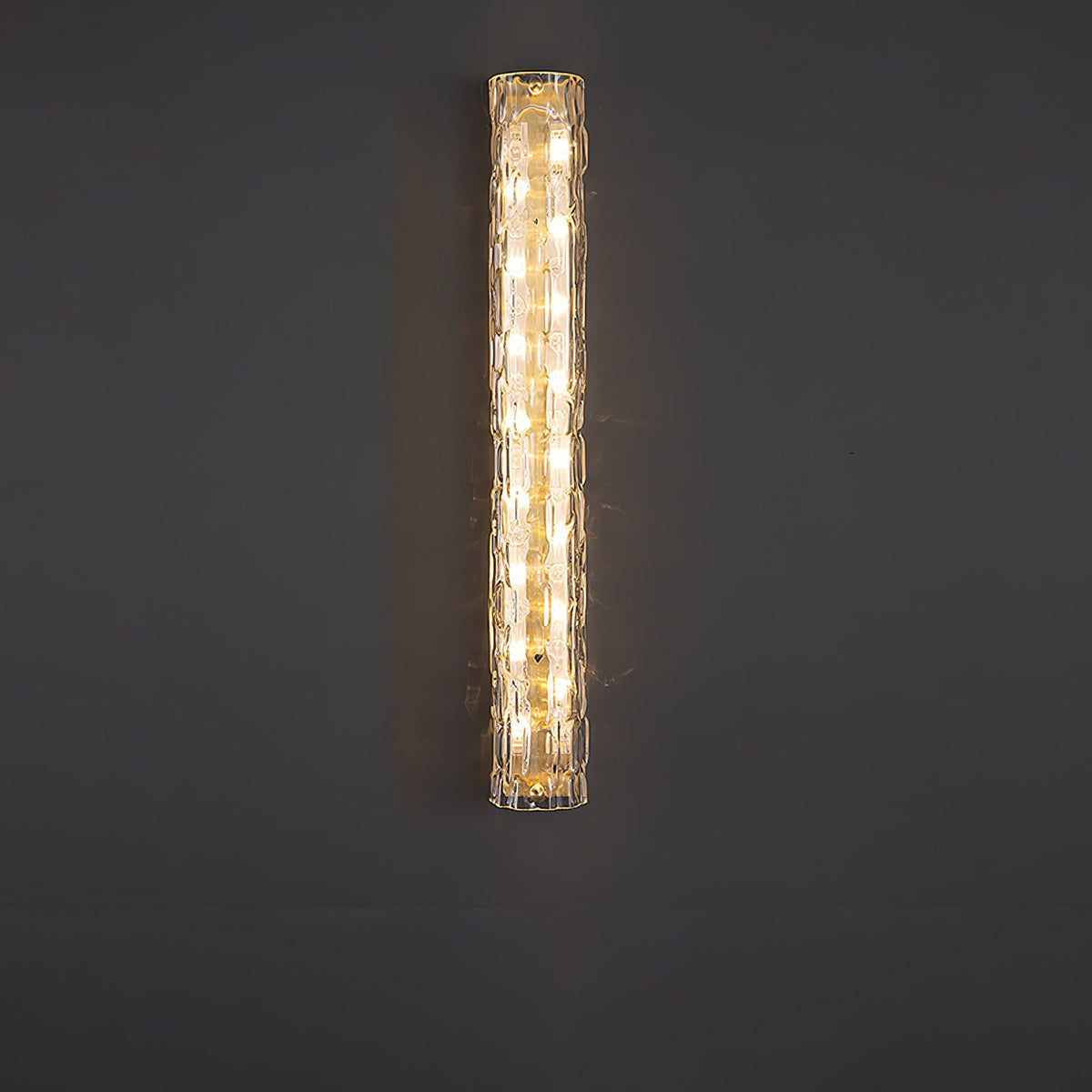 A taller Clérachic wall light glowing on a dark grey wall, its textured clear glass catching the warm light.