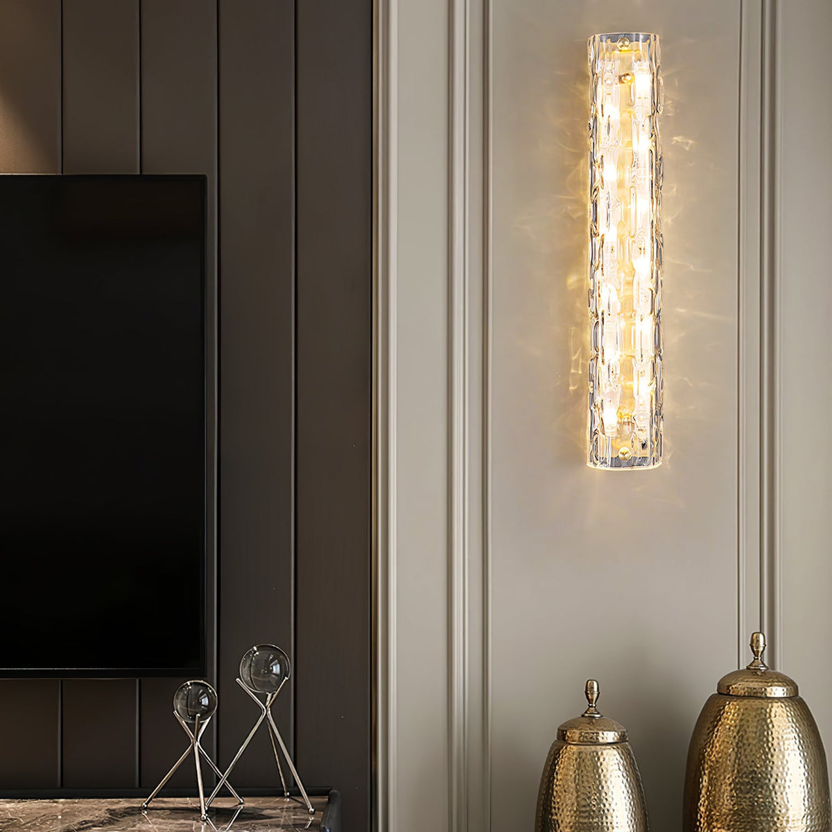 Clérachic wall sconce lit on a pale panelled wall, with a dark TV and two hammered gold urns below.