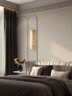 A Clérachic glass wall light lit between arched wall panels in a bedroom, with a dark curtain and a bed in grey linen.
