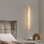 Slender Clérachic wall light lit on a pale wall above a bed with a quilted grey headboard and a glass side table.