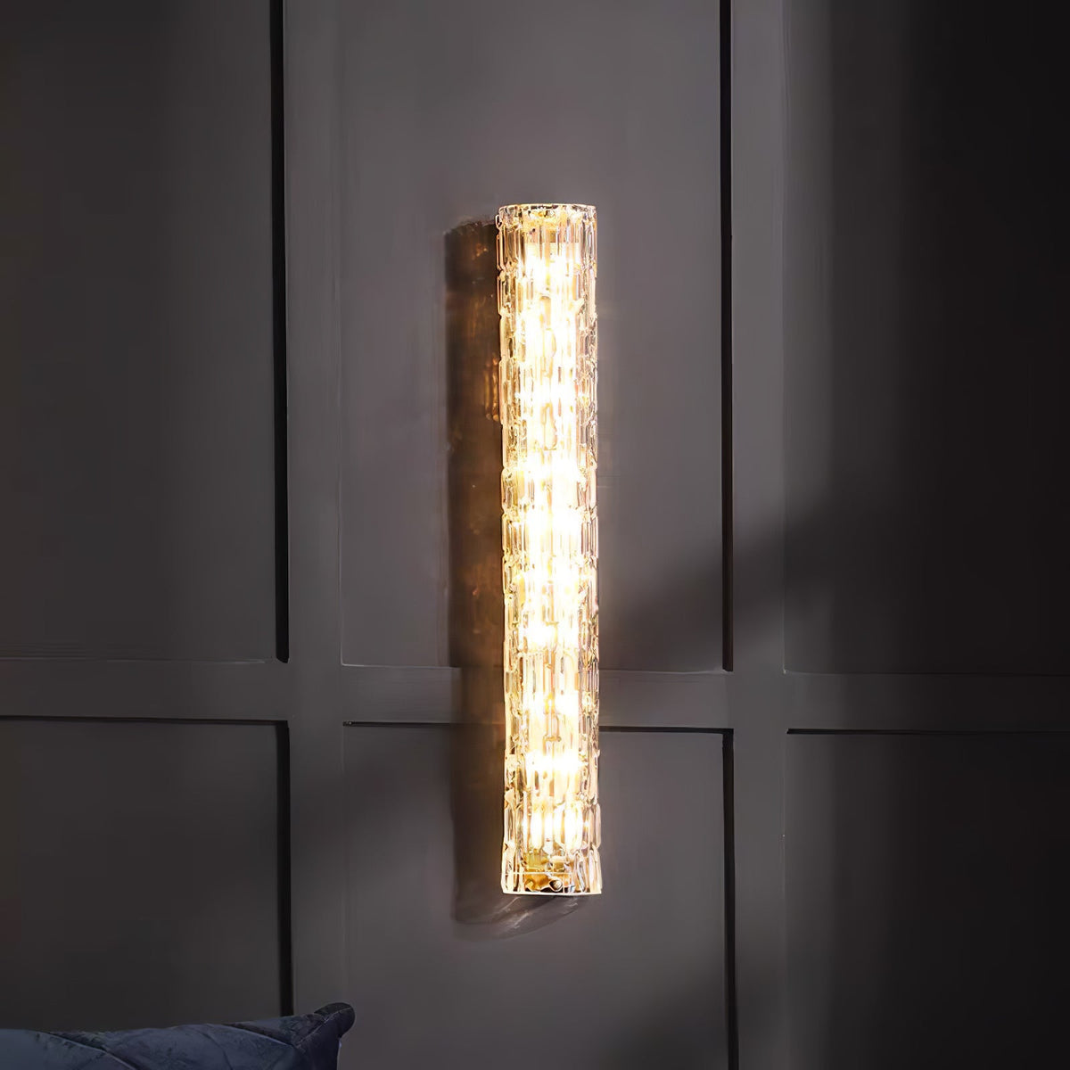 Clérachic wall light lit on a dark panelled wall, with the edge of a blue cushion at the bottom.