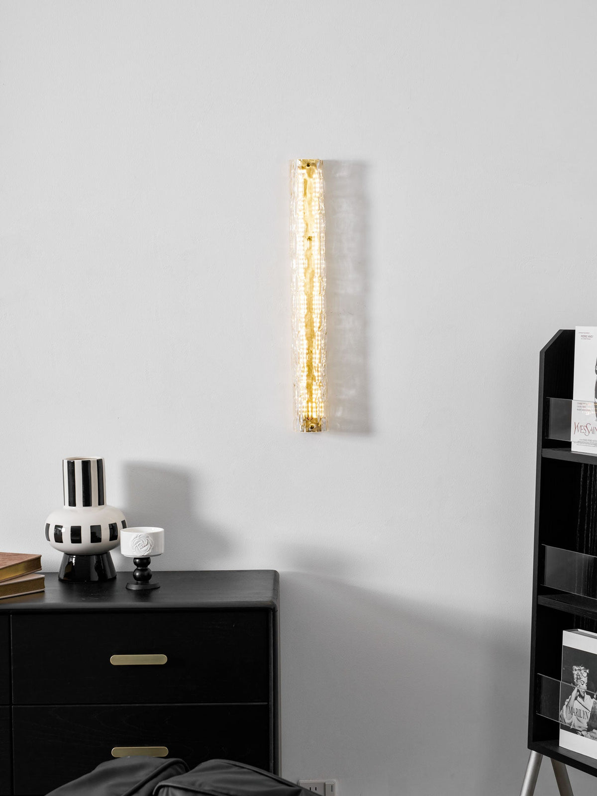 Clérachic wall light lit as a gold strip on a pale grey wall, with a black chest of drawers and a striped vase below.
