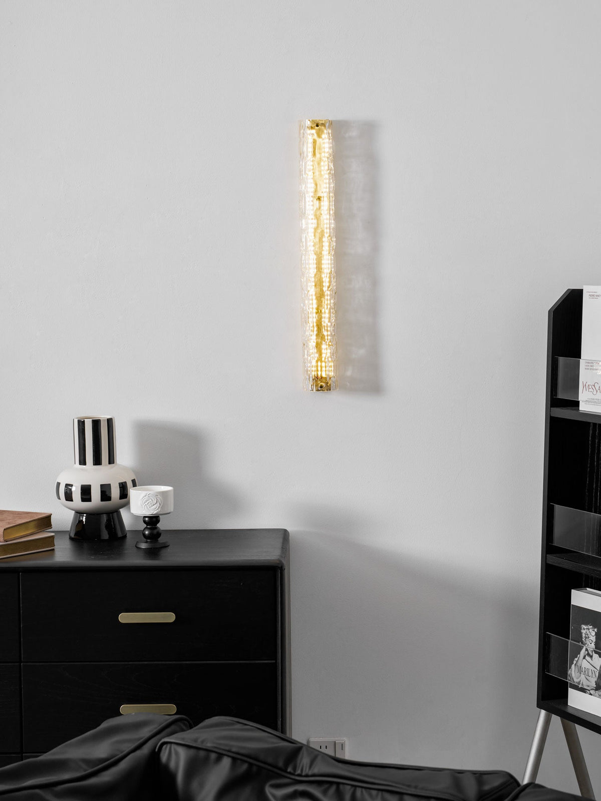 Clérachic wall light on a pale grey wall, only faintly glowing, above a black chest and a black-and-white striped vase.