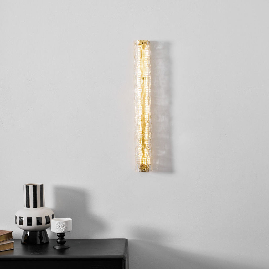 A Clérachic wall light lit warm gold on a pale wall, above a striped vase and a small white cup.