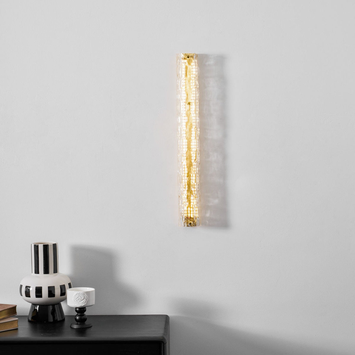 A Clérachic wall light lit warm gold on a pale wall, above a striped vase and a small white cup.