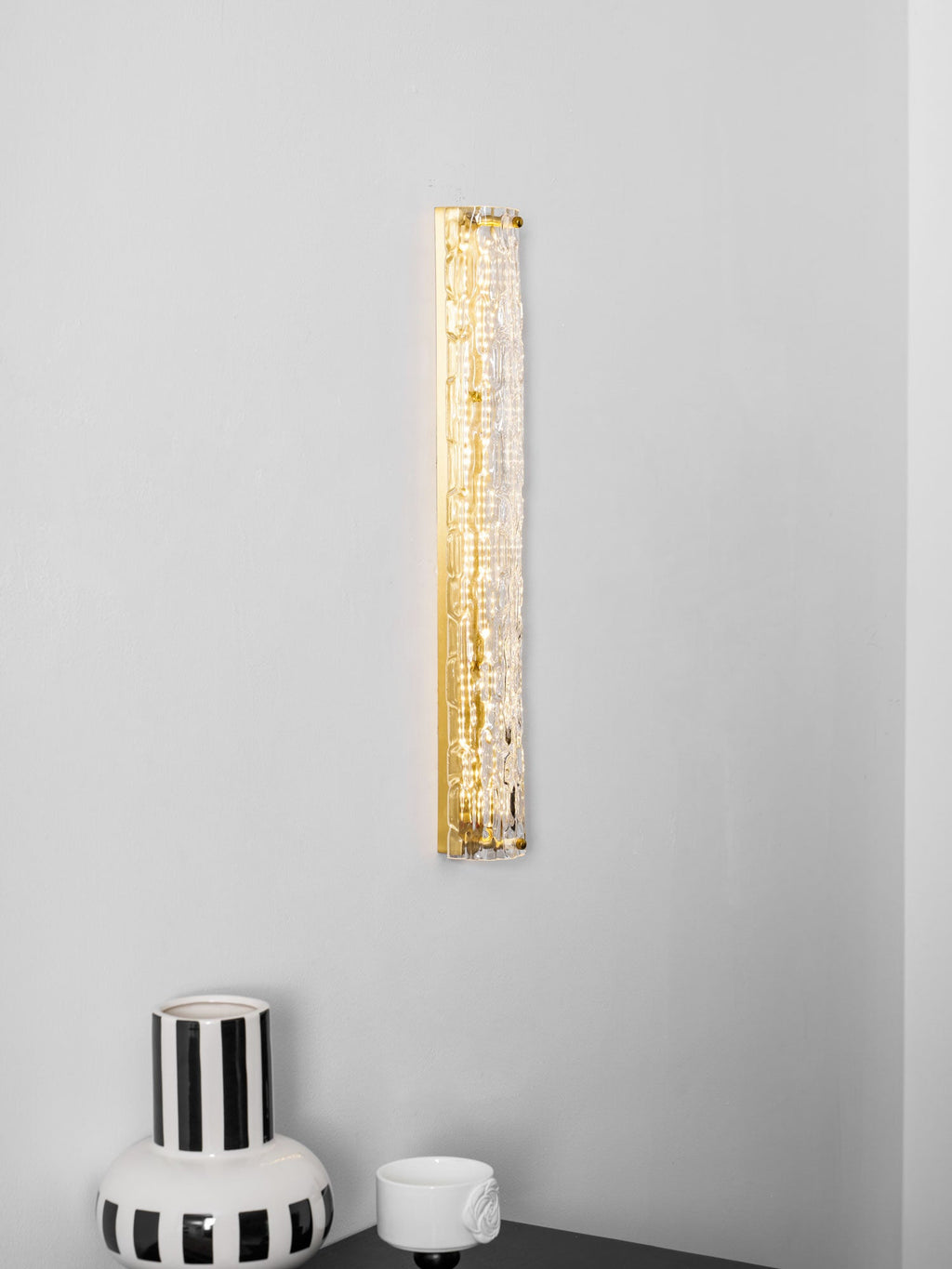Tall Clérachic wall light lit as a gold strip on a pale wall, with the rim of a striped vase at the bottom.