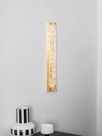Tall Clérachic wall light lit as a gold strip on a pale wall, with the rim of a striped vase at the bottom.