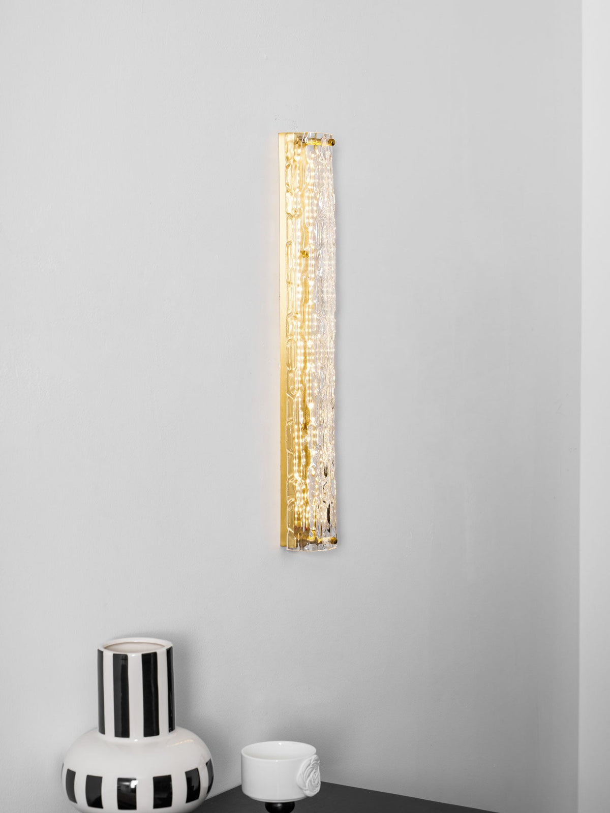 Tall Clérachic wall light lit as a gold strip on a pale wall, with the rim of a striped vase at the bottom.