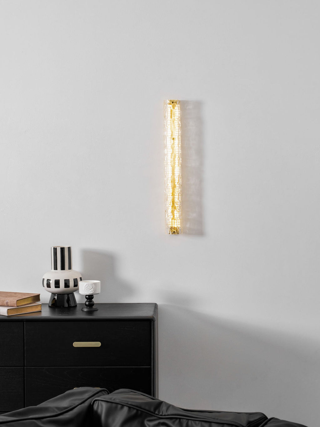 Clérachic wall light glowing softly above a black chest of drawers with brass pulls and a striped vase.