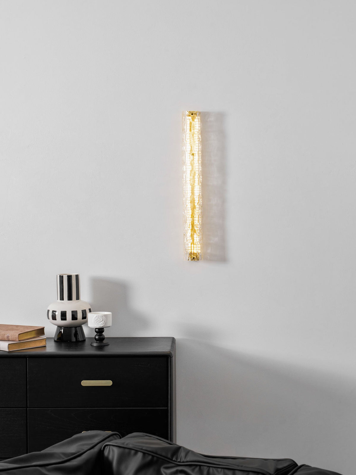 Clérachic wall light glowing softly above a black chest of drawers with brass pulls and a striped vase.