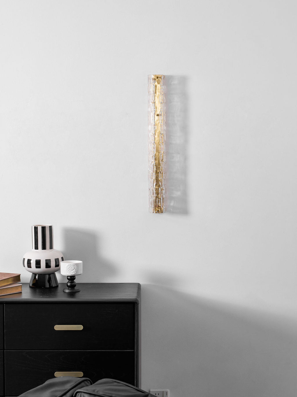 Unlit Clérachic wall light, with clear glass over a gold frame, above a black chest and a striped vase.