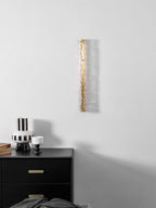 Unlit Clérachic wall light, with clear glass over a gold frame, above a black chest and a striped vase.