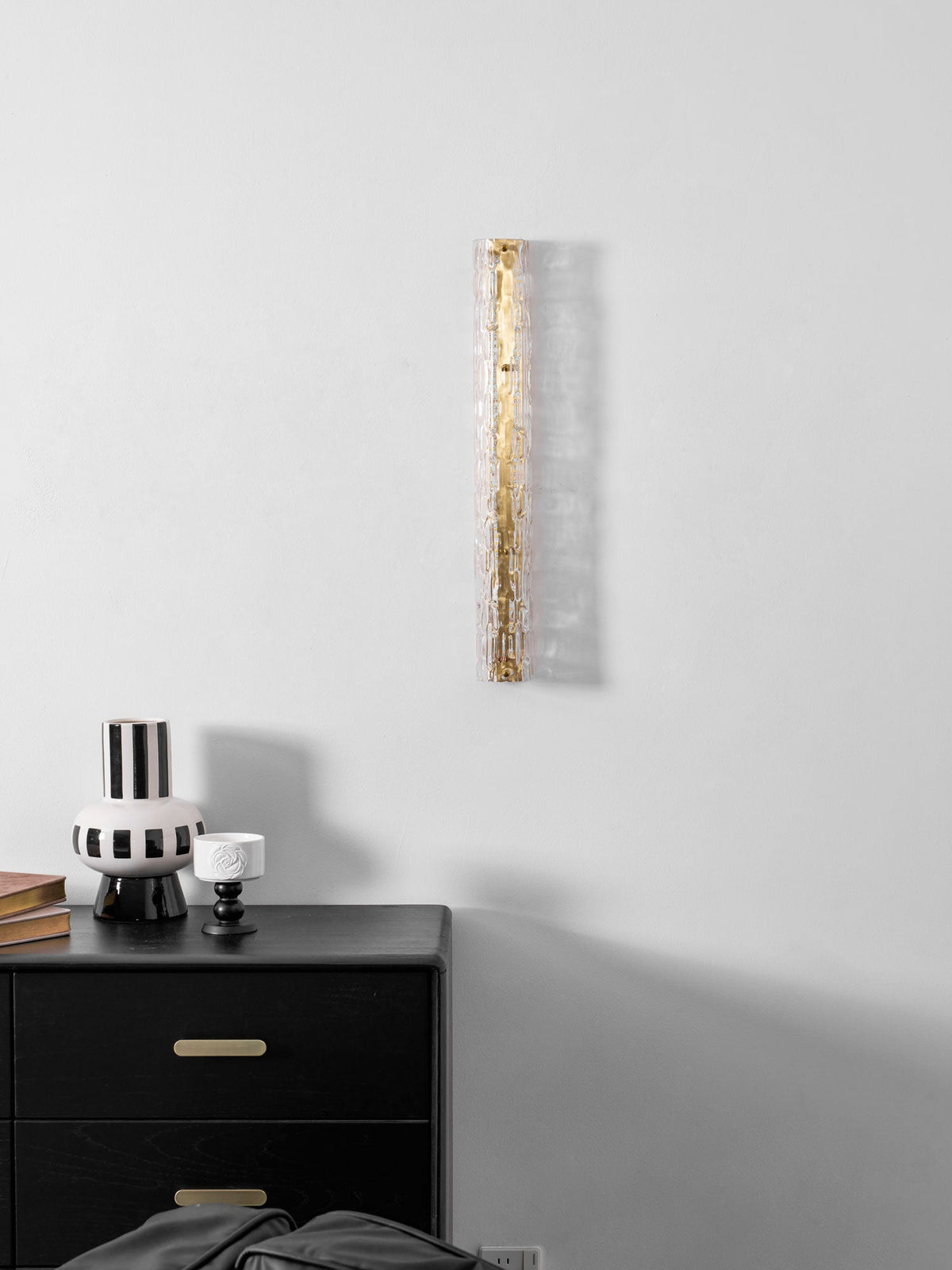 Unlit Clérachic wall light, with clear glass over a gold frame, above a black chest and a striped vase.