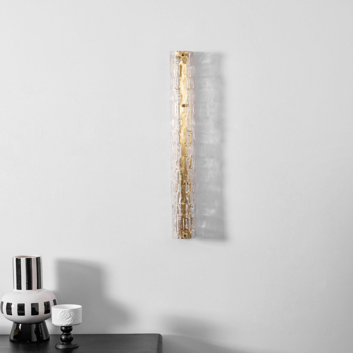 Clérachic wall light with clear rippled glass and a gold backplate, lit, with a striped vase low in the frame.