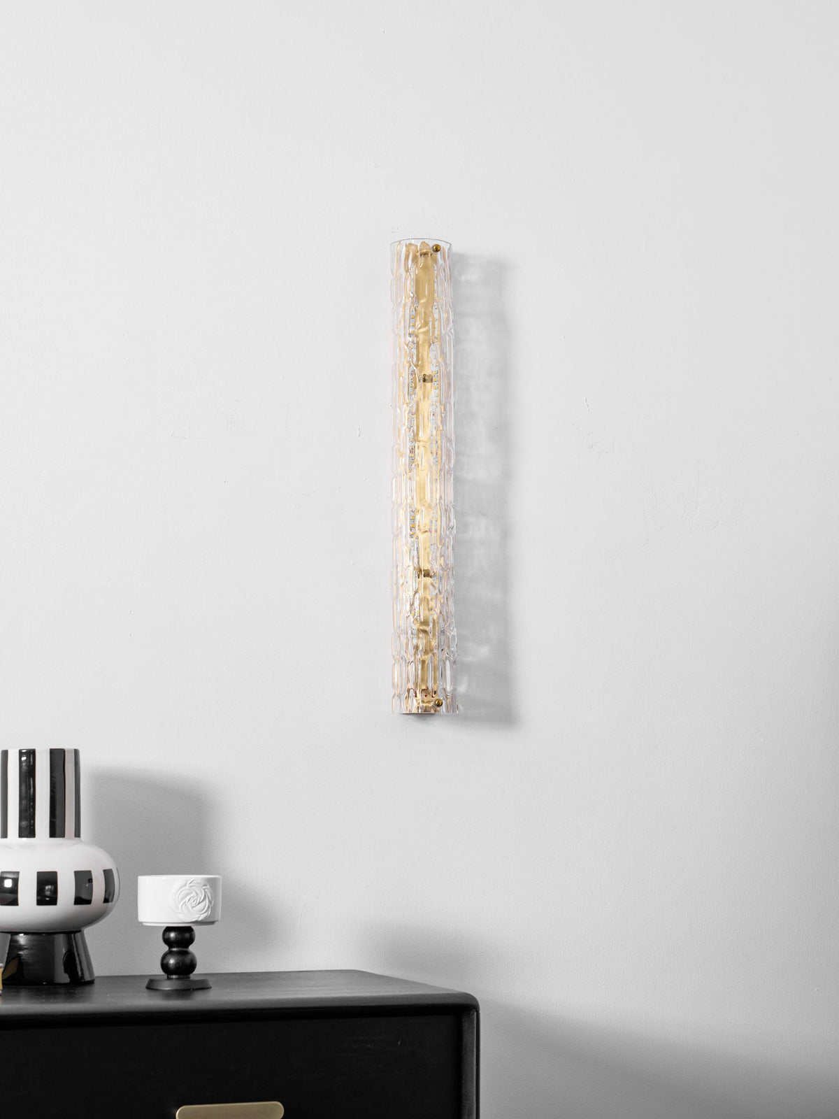 Unlit Clérachic wall light on a pale wall, the glass showing a gold backplate, with a striped vase beneath.