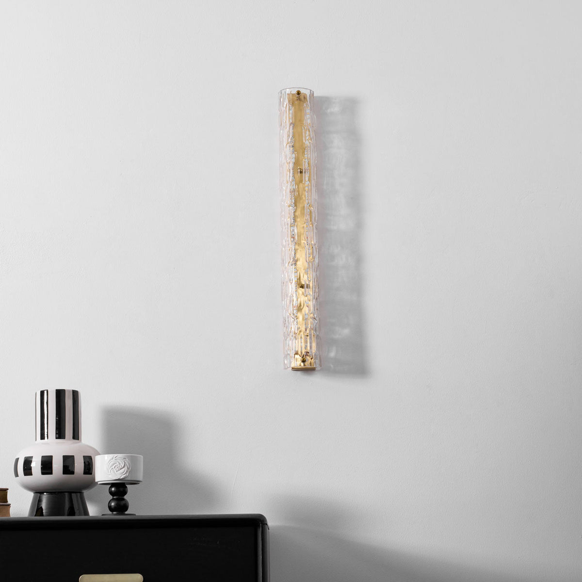 Clérachic wall light, unlit, on a pale wall above a black chest, with a striped vase and a shadow cast beside it.