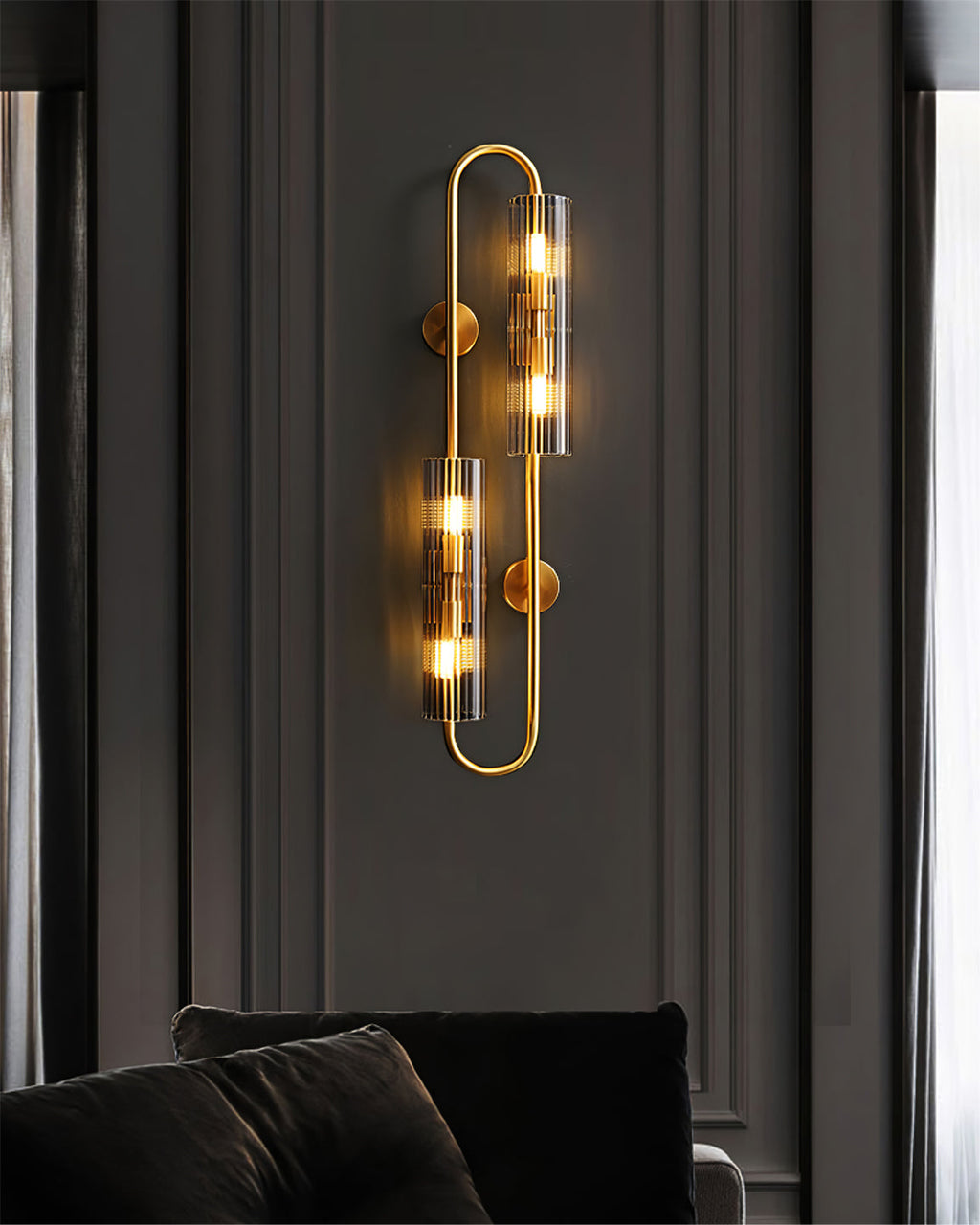 Lit on a dark panelled wall above dark cushions, this Clérachic double brass wall light has a tall looped frame and two glass tubes.