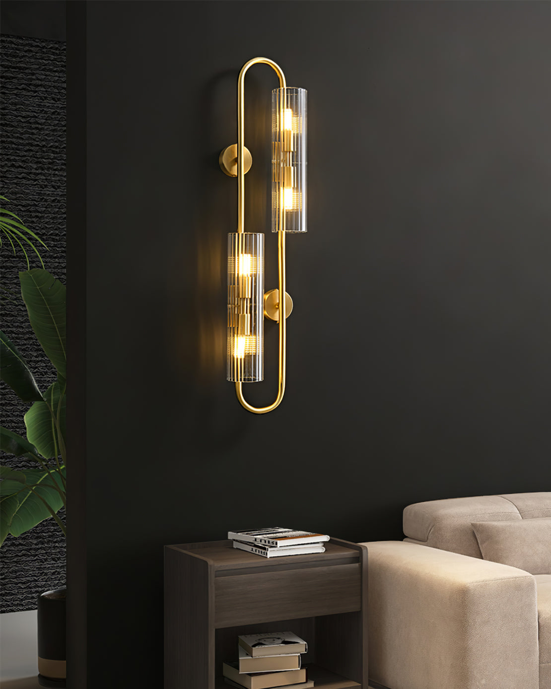 This Clérachic brass wall light glows on a dark wall above a wooden side table with books, next to a beige sofa and a trailing plant.