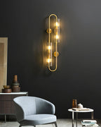 This Clérachic brass wall light lit on a dark wall above a grey armchair and a round side table, with a low sideboard to the left.