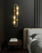 A Clérachic double brass wall light glows on a dark wall beside slatted panelling, above a nightstand and a grey bed.