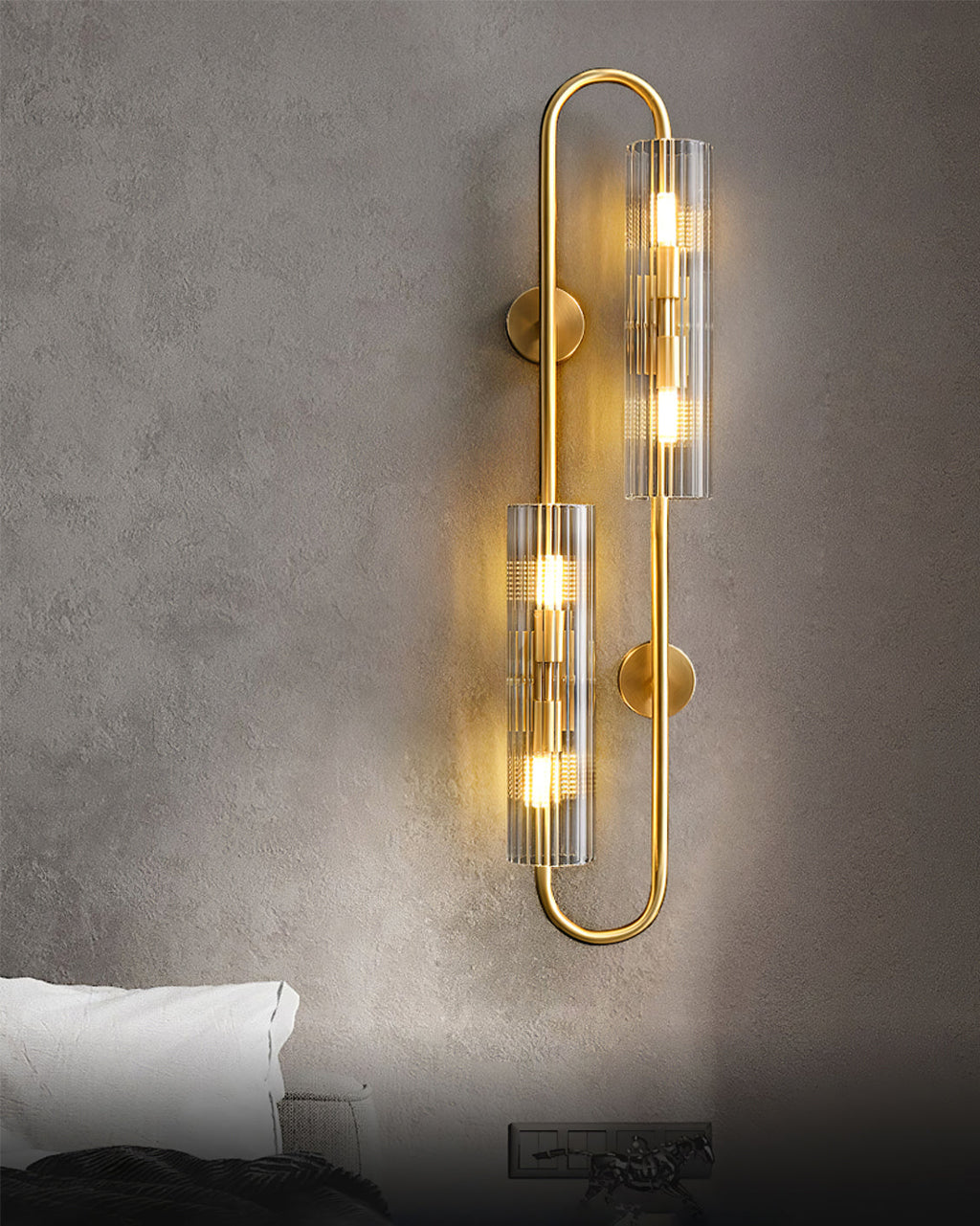 Lit on a rough grey plaster wall above a bed, this Clérachic brass wall light shows warm bulbs through its ribbed glass tubes.