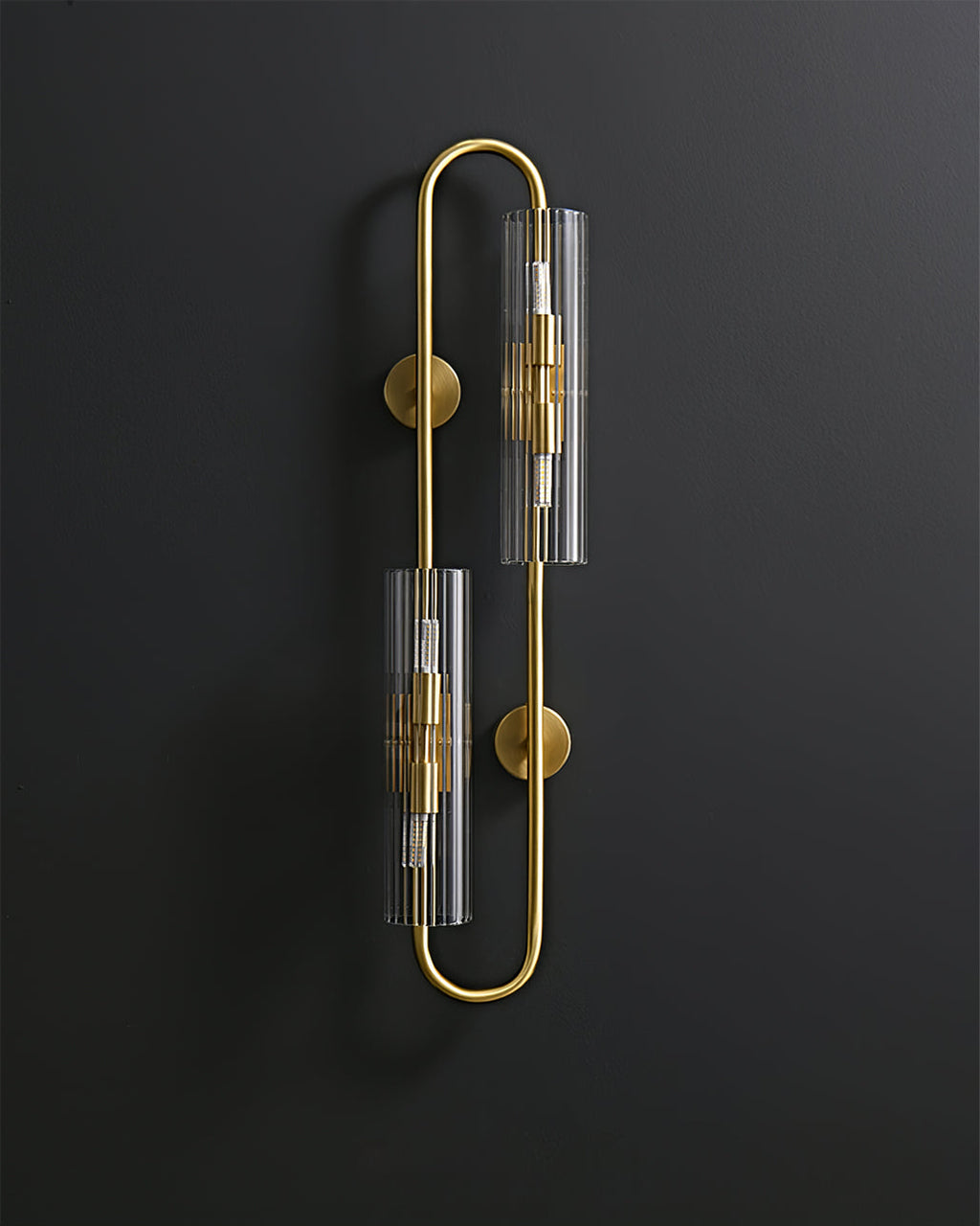 Unlit, this Clérachic brass wall light shows its two ribbed clear glass tubes and round wall plates against a dark background.
